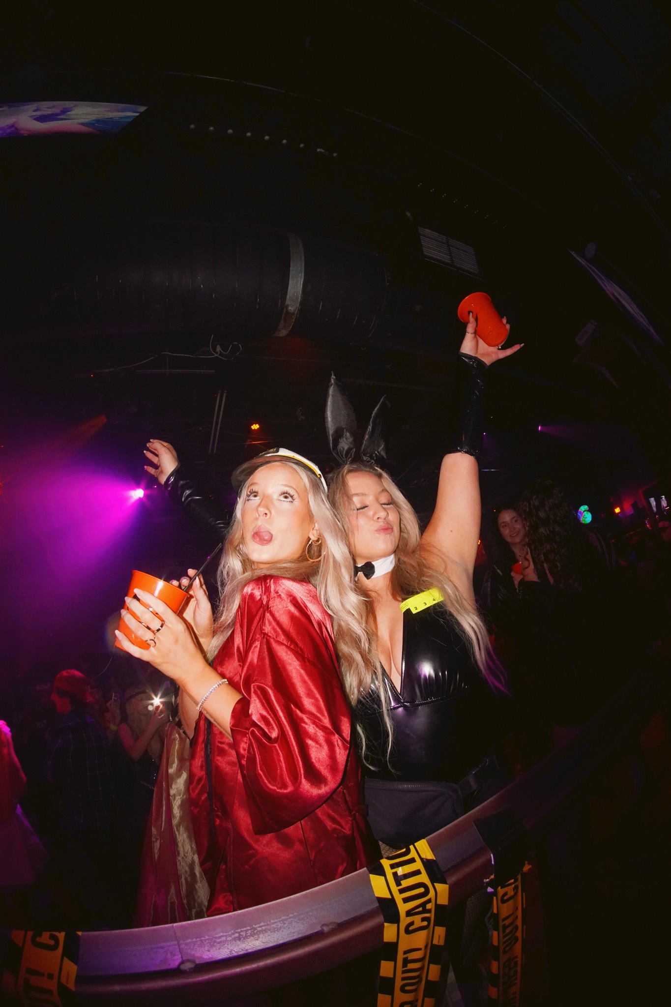 Two crawlers in costume posing back to back under purple club lighting, one in a red satin robe and sailor cap holding an orange cup, the other in black with bunny ears and an arm thrown up